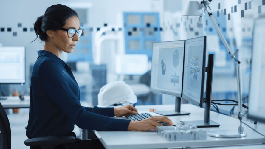 A woman sitting at a desk using a computer with 2 monitors. There is a hard hat on her desk and the background, which is blurred, looks to be a manufacturing or scince lab.