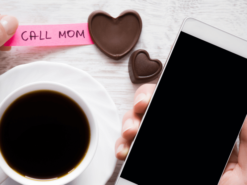 A cup of coffee and chocolate hearts on a table with a "call mom" note near one of the hearts and a person's hand holding a smart phone.