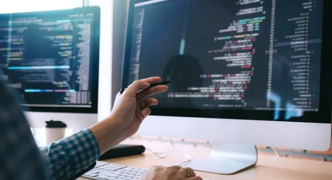 Image of computer screen containing programming code and developer's hand pointing to content.